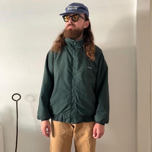 May include: A dark green zip-up jacket with a high collar, long sleeves, and elastic cuffs, paired with tan trousers, a blue baseball cap with the text Courage Bagels, and black-framed sunglasses with yellow-tinted lenses.