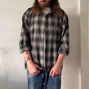 May include: A long-sleeve button-down shirt featuring a black, gray, and off-white plaid pattern. The shirt has a classic collar, a single chest pocket, and cuffs that are rolled up. It is worn over a plain white undershirt with blue denim jeans.