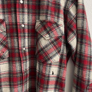 May include: A long-sleeve button-down shirt featuring a red, grey, and black plaid pattern. The shirt includes two chest pockets with buttoned flaps, silver-toned snap buttons down the front, and a pointed western-style yoke across the shoulders.