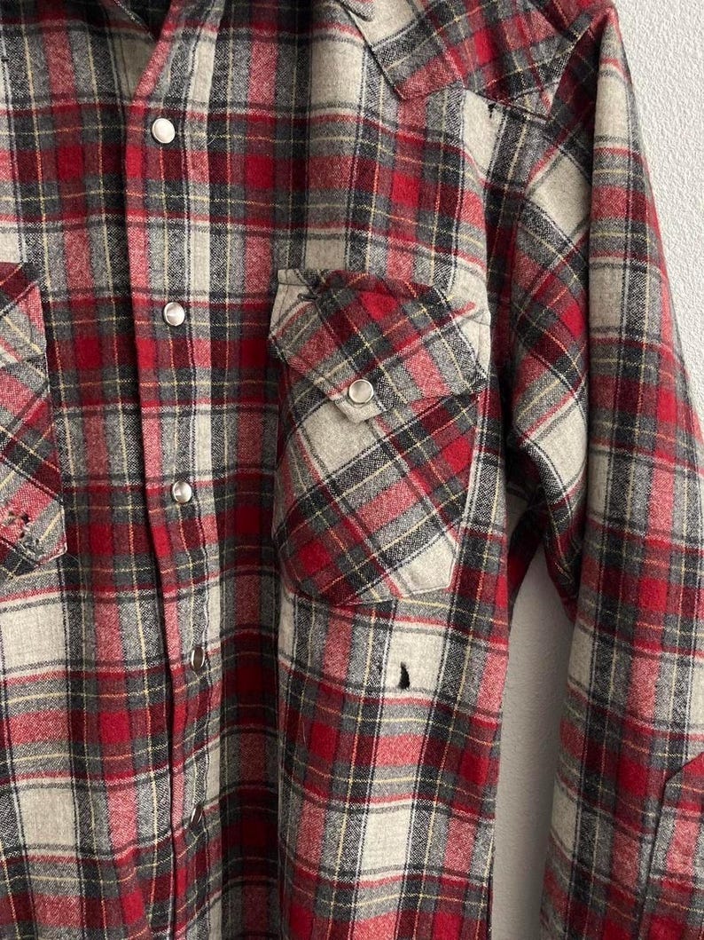 May include: A long-sleeve button-down shirt featuring a red, grey, and black plaid pattern. The shirt includes two chest pockets with buttoned flaps, silver-toned snap buttons down the front, and a pointed western-style yoke across the shoulders.