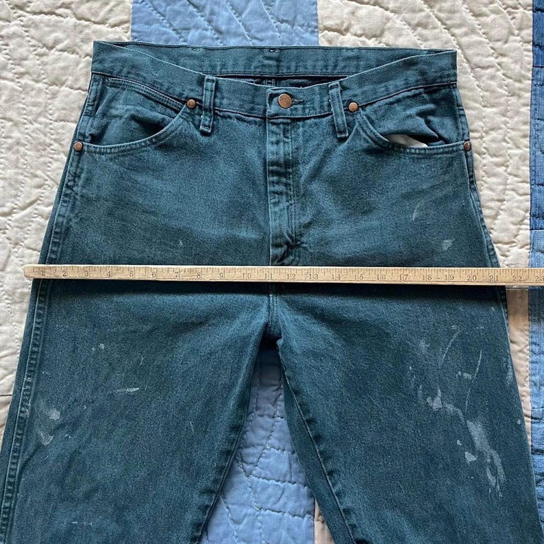 Puede incluir: Vaqueros de mezclilla en color verde azulado oscuro con dise&ntilde;o de cintura alta, cierre de bot&oacute;n y cremallera, remaches de color cobre y salpicaduras de pintura clara. Una regla de madera mide horizontalmente 56 cm de ancho en la cintura.