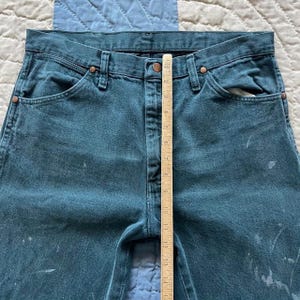 Puede incluir: Vaqueros de mezclilla en color verde azulado oscuro con cierre de botones, remaches de tono cobre y salpicaduras de pintura clara. Una regla de madera est&aacute; colocada verticalmente en el centro, midiendo unos 35,5 cm desde la cintura.