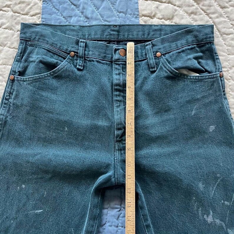 Puede incluir: Vaqueros de mezclilla en color verde azulado oscuro con cierre de botones, remaches de tono cobre y salpicaduras de pintura clara. Una regla de madera est&aacute; colocada verticalmente en el centro, midiendo unos 35,5 cm desde la cintura.