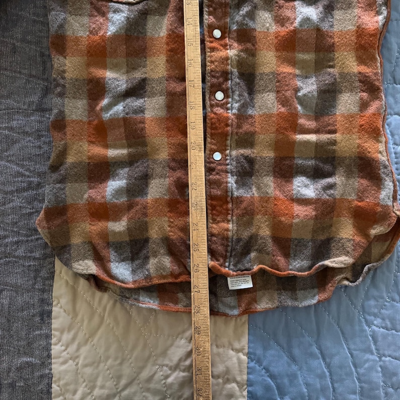 K&ouml;nnte beinhalten: Ein lang&auml;rmeliges Hemd mit Knopfleiste und Karomuster in Orange-, Braun- und Graut&ouml;nen. Ein Holzlineal liegt vertikal &uuml;ber der Mitte und zeigt eine L&auml;ngenmessung von etwa 35 cm bis 81 cm an.