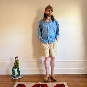 May include: A light blue denim button-down shirt, beige knee-length shorts, a brown knit beanie, and tan slip-on shoes. A small figurine of a person in a yellow shirt and green pants stands on the floor near the feet of the main subject.