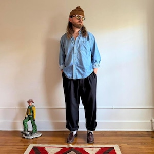 May include: A light blue long-sleeve button-down shirt, black trousers with a cuffed hem, brown leather loafers, a brown knit beanie, and sunglasses. A small figurine of a person in a yellow shirt and green pants stands on the floor.