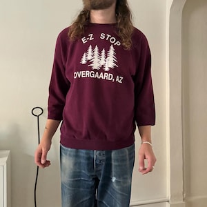 May include: A maroon crewneck sweatshirt with three-quarter length sleeves. The front features white screen-printed text that reads E-Z STOP and OVERGAARD, AZ, with a graphic of a cluster of pine trees positioned between the two lines of text.