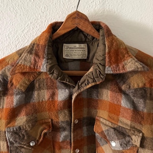 K&ouml;nnte beinhalten: Ein lang&auml;rmeliges Pendleton-Hemd mit einem Karomuster in Orange, Braun und Grau. Das Hemd hat einen spitzen Kragen, zwei Brusttaschen mit Druckkn&ouml;pfen und ein Etikett mit der Aufschrift Pendleton High Grade Western Wear Portland Oregon 100% Virgin Wool L.