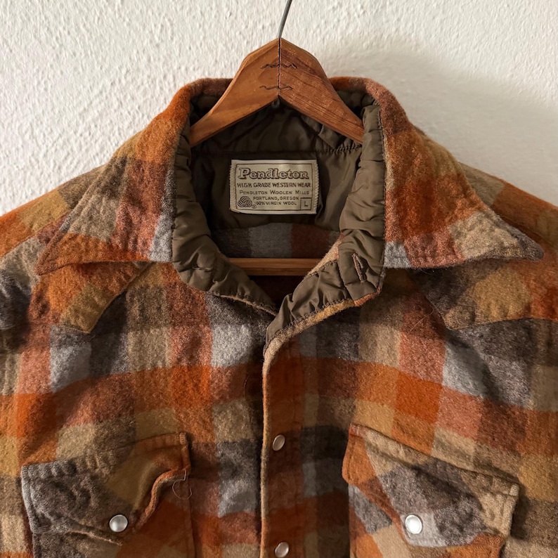 K&ouml;nnte beinhalten: Ein lang&auml;rmeliges Pendleton-Hemd mit einem Karomuster in Orange, Braun und Grau. Das Hemd hat einen spitzen Kragen, zwei Brusttaschen mit Druckkn&ouml;pfen und ein Etikett mit der Aufschrift Pendleton High Grade Western Wear Portland Oregon 100% Virgin Wool L.