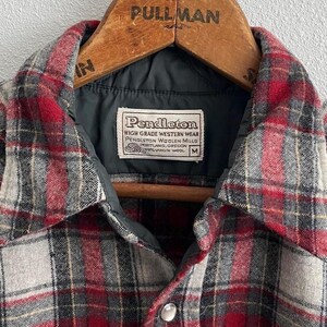 May include: A red, grey, and black plaid wool shirt with a dark grey quilted lining and a white label reading Pendleton High Grade Western Wear Pendleton Woolen Mills Portland Oregon 100% Virgin Wool M, hanging on a wooden Pullman hanger.