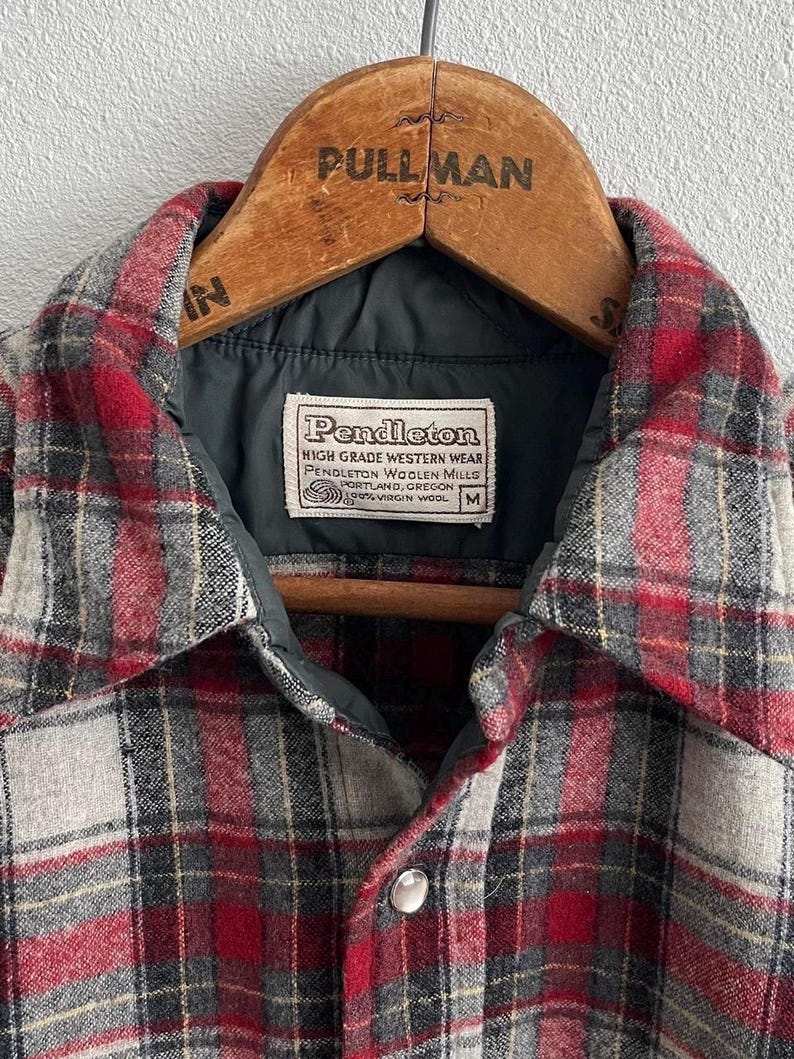 May include: A red, grey, and black plaid wool shirt with a dark grey quilted lining and a white label reading Pendleton High Grade Western Wear Pendleton Woolen Mills Portland Oregon 100% Virgin Wool M, hanging on a wooden Pullman hanger.