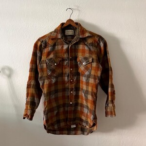 K&ouml;nnte beinhalten: Ein lang&auml;rmeliges Hemd mit Knopfleiste und einem Karomuster in den Farben gebranntes Orange, Braun und Grau. Das Hemd verf&uuml;gt &uuml;ber einen spitzen Kragen, zwei Brusttaschen mit Druckknopfverschluss und eine Knopfleiste mit metallischen Druckkn&ouml;pfen.