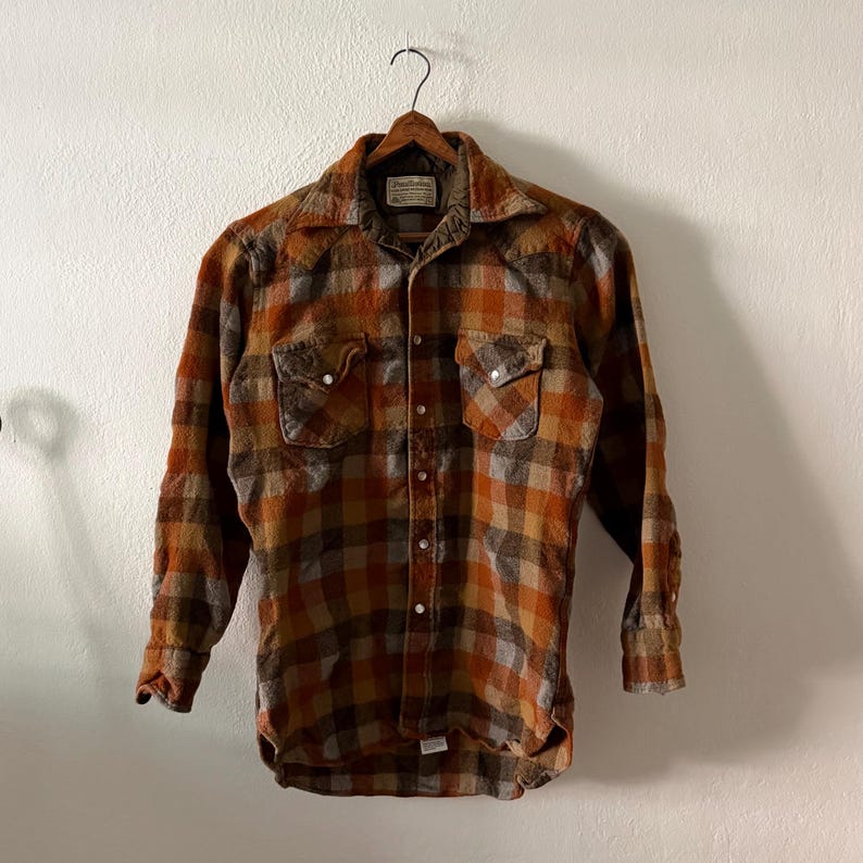 K&ouml;nnte beinhalten: Ein lang&auml;rmeliges Hemd mit Knopfleiste und einem Karomuster in den Farben gebranntes Orange, Braun und Grau. Das Hemd verf&uuml;gt &uuml;ber einen spitzen Kragen, zwei Brusttaschen mit Druckknopfverschluss und eine Knopfleiste mit metallischen Druckkn&ouml;pfen.