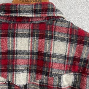 May include: A long-sleeve button-up shirt featuring a red, grey, black, and white plaid pattern. The shirt has a pointed collar and a western-style yoke across the back shoulders. The fabric has a soft, brushed texture throughout the garment.