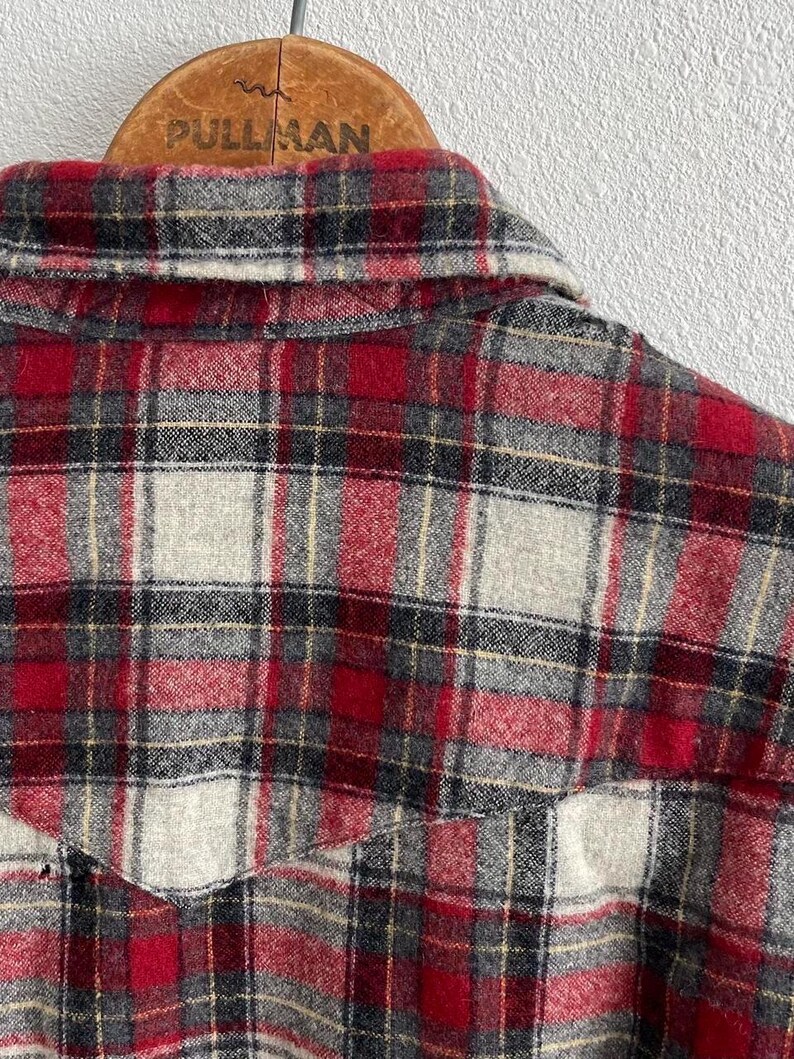 May include: A long-sleeve button-up shirt featuring a red, grey, black, and white plaid pattern. The shirt has a pointed collar and a western-style yoke across the back shoulders. The fabric has a soft, brushed texture throughout the garment.