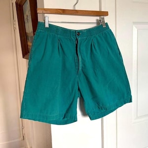 May include: Teal green shorts featuring an elastic waistband, a front button closure, and a zippered fly. The shorts have two front pleats and side pockets, hanging from a wooden hanger against a plain white door background.