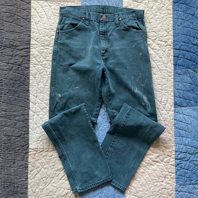 Puede incluir: Unos vaqueros de mezclilla color verde azulado con dise&ntilde;o de cintura alta, cierre de bot&oacute;n y cremallera, cinco bolsillos y remaches de color cobre. La tela muestra salpicaduras de pintura clara y marcas de desgaste por toda la prenda.
