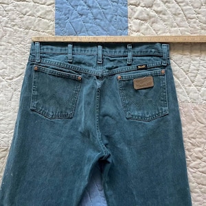 Puede incluir: Vaqueros de mezclilla color verde azulado con canes&uacute; trasero, dos bolsillos traseros con costura en W, remaches de color cobre y un parche de cuero marr&oacute;n de Wrangler en el bolsillo derecho. Una regla de madera muestra una anchura de 48 cm.