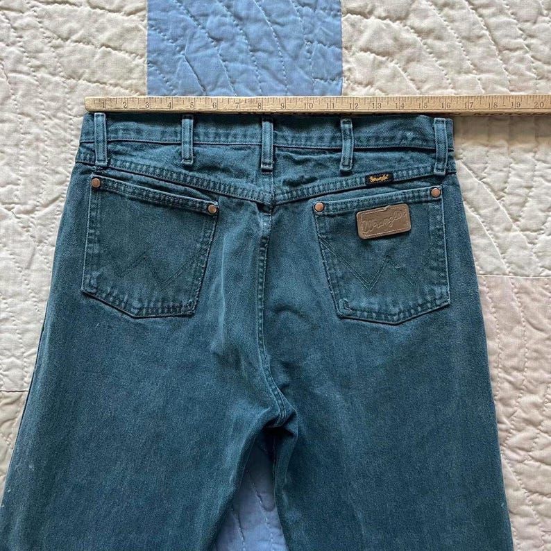 Puede incluir: Vaqueros de mezclilla color verde azulado con canes&uacute; trasero, dos bolsillos traseros con costura en W, remaches de color cobre y un parche de cuero marr&oacute;n de Wrangler en el bolsillo derecho. Una regla de madera muestra una anchura de 48 cm.