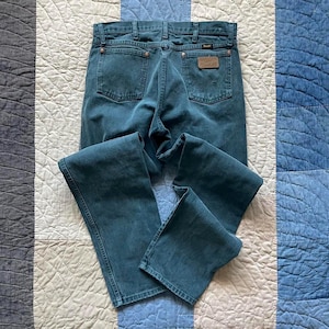 Puede incluir: Pantalones vaqueros de color verde azulado con un dise&ntilde;o cl&aacute;sico de cinco bolsillos, cierre de botones y remaches de color cobre. Los bolsillos traseros muestran la costura caracter&iacute;stica de Wrangler y un parche de cuero marr&oacute;n.