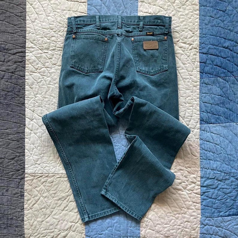 Puede incluir: Pantalones vaqueros de color verde azulado con un dise&ntilde;o cl&aacute;sico de cinco bolsillos, cierre de botones y remaches de color cobre. Los bolsillos traseros muestran la costura caracter&iacute;stica de Wrangler y un parche de cuero marr&oacute;n.