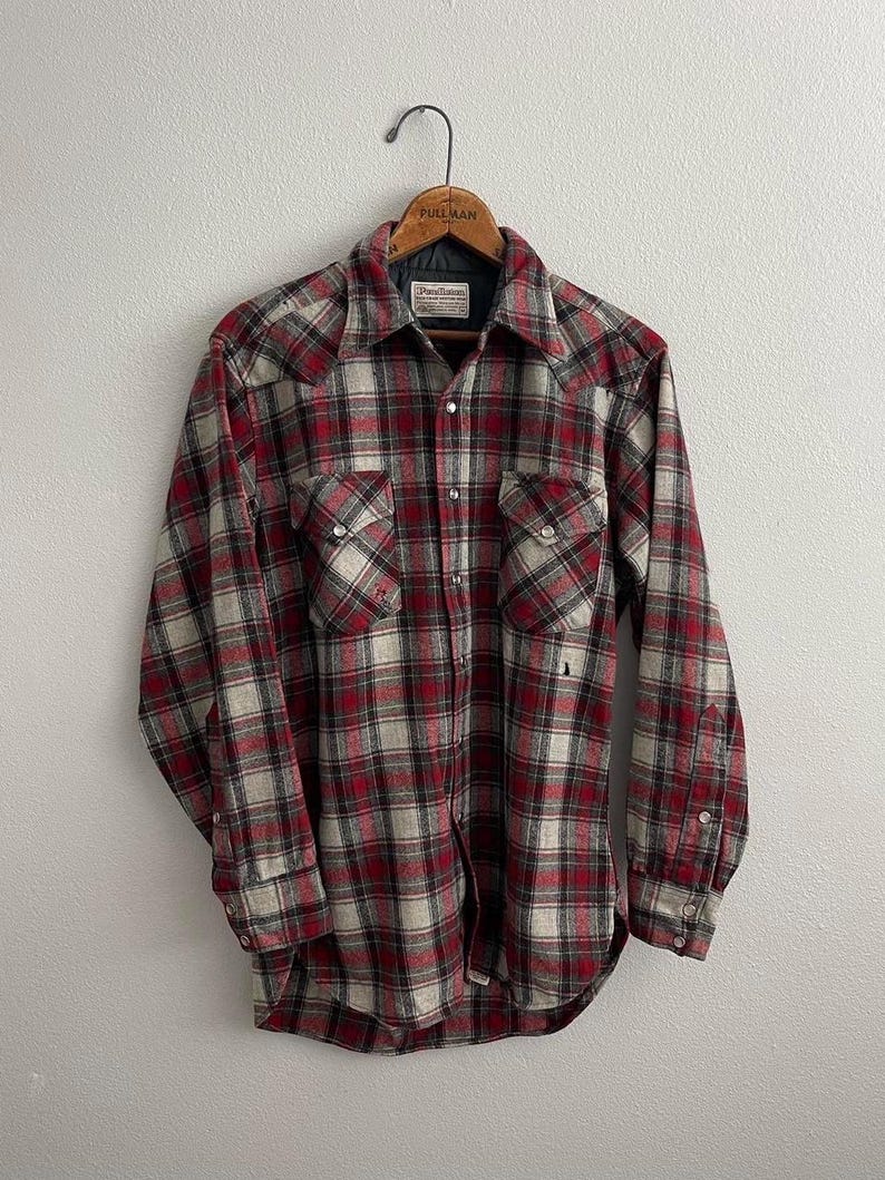 May include: A long-sleeve button-down shirt featuring a red, black, and off-white plaid pattern. The shirt includes two chest pockets with button-down flaps, a pointed collar, and silver-toned snap buttons down the front and on the cuffs.