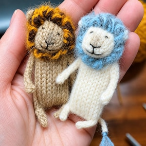 May include: Two small, hand-knitted lion figurines. One lion has a tan body with a golden-brown and dark brown mane. The other lion has a cream-colored body with a light blue mane. Both have embroidered facial features and small tails.