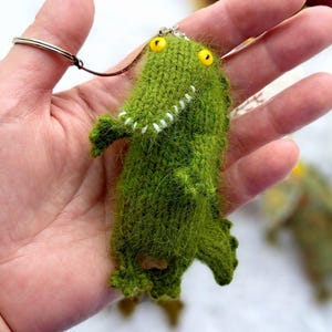 May include: A handmade green knitted crocodile keyring with yellow eyes and white teeth. The small plush alligator has a silver keyring attached. The crocodile is standing upright with its arms out. The keyring is held in a person's hand.