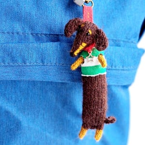 May include: A knitted brown dachshund dog keyring with yellow paws and a green and white striped top. The dog has a red collar and is attached to a red strap with a silver clasp. The background is a blue fabric.
