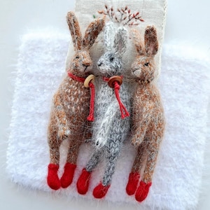 May include: Three knitted rabbit figurines. Two are brown and one is gray, each with red boots and a red string collar with a wooden button. They stand on a white, fluffy surface, with a beige embroidered backdrop.