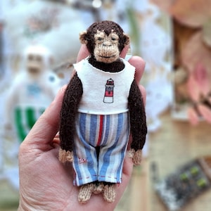 Op de afbeelding: Een handgemaakte miniatuur aap pop met bruine vacht, gekleed in een wit shirt met een vuurtoren grafisch en blauw gestreepte broek met een rode streep. De pop is gemaakt van garen en heeft een gedetailleerd gezicht.