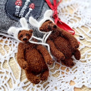 Könnte beinhalten: Zwei Miniatur-Teddybären in Braun mit weißen Hasenmützen. Die Bären sind gestrickt und haben eine weiße Schnur um den Hals. Im Hintergrund befindet sich ein grauer Stoff mit einem Pilzdesign, zusammen mit einem roten Band und einer weißen Spitzendecke.
