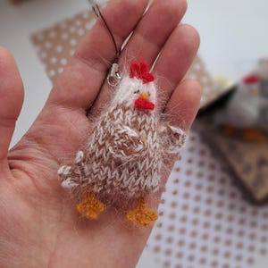 May include: A miniature knitted chicken charm, featuring a red comb and wattle, a white head, and a brown and white body. The chicken has yellow feet and a small silver clasp. The charm is held in a hand.