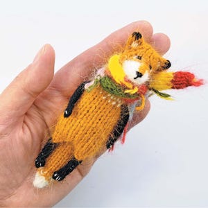 以下が含まれることがあります： オレンジ、黒、白、黄色、緑、赤の色合いの手編みの小さなキツネの置物。キツネはカラフルなスカーフを身につけ、黒い足を持っています。キツネは横になっています。置物は人の手で持たれています。