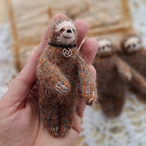 Könnte beinhalten: Handgefertigte Miniatur-Faultier-Puppe mit braun-grauem Strickfell. Das Faultier trägt einen schwarzen Kragen mit einem goldfarbenen Zahnrad-Anhänger. Im Hintergrund sind zwei weitere Faultier-Puppen zu sehen, was ein handwerkliches Thema erzeugt.