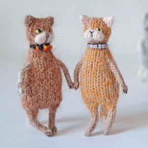 Può includere: Due figurine di gatti lavorate a maglia a mano che si tengono per mano. Un gatto è marrone con un collare arancione e nero, l'altro è arancione con un collare bianco. I gatti hanno piccoli dettagli e sono in piedi.