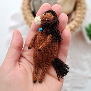 May include: A handmade brown knitted horse figurine with a black mane and tail. The horse has a white muzzle and a small blue bead around its neck. The figurine is approximately 7.6 cm tall.