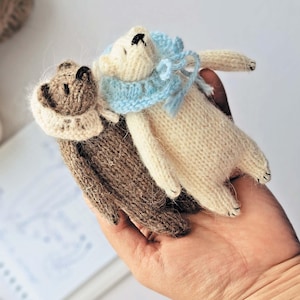 May include: Two handmade knitted teddy bears. One is brown with a cream-colored collar, and the other is off-white with a light blue collar. The bears are lying side by side, showcasing their soft, textured appearance. The background is a neutral color.