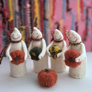 Könnte beinhalten: Vier handgefertigte, cremefarbene, gestrickte Geisterfiguren mit lächelnden Gesichtern, die kleine Kürbisse in verschiedenen Farben halten. Jeder Geist trägt einen bunten Schal. Vor den Figuren sitzt ein einzelner orangefarbener Kürbis.