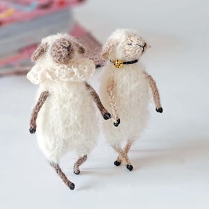 Op de afbeelding: Twee handgemaakte, pluizige witte schaapjes met bruine accenten. Eén schaap heeft een gekruld kraagje, de ander een zwarte kraag met een gouden bel. De schapen hebben bruine poten en zwarte hoeven, op een wit oppervlak.