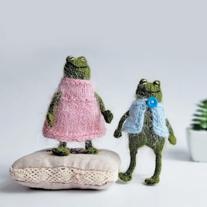 May include: Two handmade green frog figurines. One frog wears a pink knitted dress and stands on a small cushion. The other frog wears a blue knitted waistcoat with a blue button. A whimsical craft.