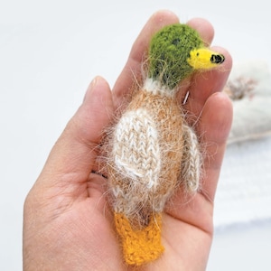 Peut inclure: Figurine de caneton miniature fait à la main. Le caneton a une tête verte, un bec jaune et des pieds orange. Le corps est un mélange de laine tricotée marron et blanche, créant un aspect doux et texturé. Un article charmant et artisanal.
