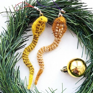 Pode incluir: Dois enfeites de cobra feitos à mão, tricotados em amarelo e castanho, com fechos prateados e fios para pendurar. Os enfeites são exibidos em uma coroa verde com uma bola dourada e um pinha.