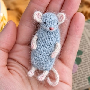 May include: A small, hand-knitted mouse figurine featuring a fuzzy, light blue body with contrasting cream-colored ears, arms, legs, and a curled tail. The mouse has tiny black eyes and a small pink nose, crafted with a soft, textured yarn finish.