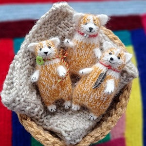 Puede incluir: Tres figuras de perros corgi tejidas a mano en una cesta tejida. Los corgis tienen pelaje naranja y blanco, con pequeños ojos y narices negros. Cada corgi tiene un collar único, uno con una flor verde, uno con un collar rojo y otro con un colgante de pluma.