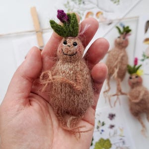 May include: A knitted mandrake doll with a smiling face, green leafy hair, and a purple flower. The doll is held in a hand, with two other similar dolls in the background. The dolls are a light brown colour.