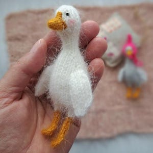 Pode incluir: Um ganso branco feito à mão, tricotado, com bico e patas laranja. O ganso tem uma textura fofa e é segurado em uma mão. Um segundo ganso, menor, é visível ao fundo.