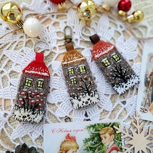 May include: Three handmade house-shaped keychains with red, brown, and white knitted details. Each keychain features a house design with glowing yellow windows, a snowy roof, and a decorative tree or floral element. The keychains are displayed with Christmas ornaments.