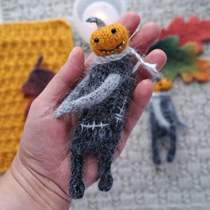 May include: Handmade Halloween-themed plush doll. The doll features a smiling orange pumpkin head, gray arms, and a dark gray body with white stitching details. The doll is held in a hand, with a second doll visible in the background. The overall theme is Halloween.