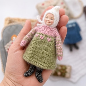 May include: A small doll with a painted face wearing a white knitted bonnet, a pink and olive green sweater with small heart patterns, and dark gray knitted leggings. The doll is crafted with a soft, textured yarn finish and features a detailed face.